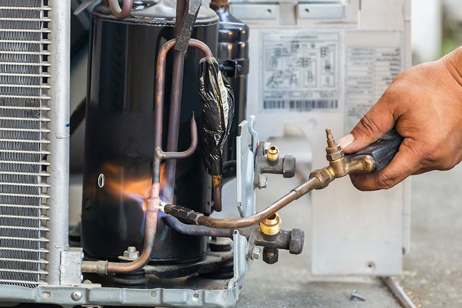 HVAC technician brazing copper refrigerant lines on an air conditioner compressor with a torch